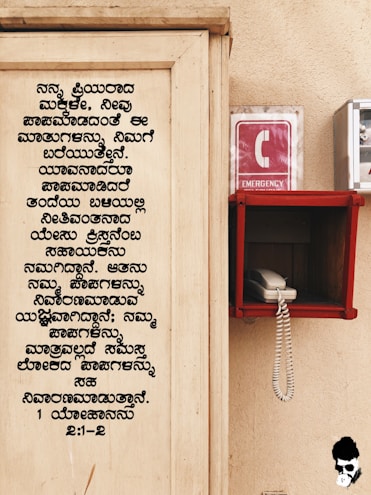 A wooden board with text written in Kannada is on the left side. On the right, there is an emergency phone booth with a red sign displaying a white 'C' and the word 'EMERGENCY'. A white corded phone is placed inside a red holder mounted on the wall.