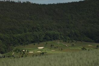 A lush green golf course is surrounded by dense forested hills. The fairways are neatly maintained, and several sand bunkers are visible across the course. The scene is peaceful, with no visible people or activity.