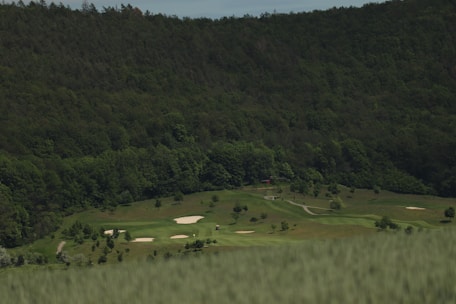 A lush green golf course is surrounded by dense forested hills. The fairways are neatly maintained, and several sand bunkers are visible across the course. The scene is peaceful, with no visible people or activity.