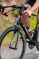 A cyclist grips the handlebars of a black road bicycle with sleek design. The bicycle is equipped with a small black light and a Garmin device mounted on the handlebars. The cyclist wears a black and red cycling outfit and is riding on a path bordered by blurred greenery, indicating motion.