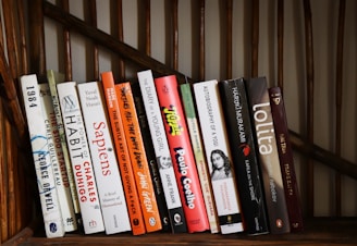 A collection of books is neatly arranged on a wooden shelf, each displaying a variety of colorful spines featuring titles from different genres and authors.