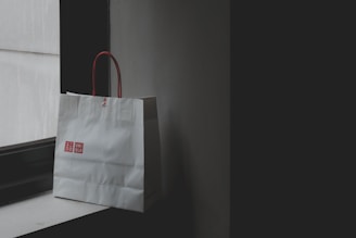 A Uniqlo shopping bag with red handles sits on a windowsill in a dimly lit room. The lighting creates a contrast between the white bag and the dark surroundings.