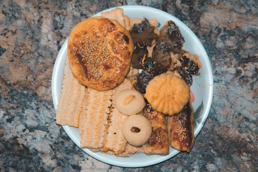 A plate filled with a variety of baked goods, including cookies, crackers, and pastries. The items have different shapes and textures, with some topped with sesame seeds or raisins. The background is a textured stone surface, which contrasts with the white plate.