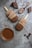 Two chocolate-covered ice cream bars with wooden sticks lie on a textured gray surface. They are surrounded by chocolate drizzle and splatters, a spoon with melted chocolate, and several pieces of broken chocolate. A small glass cup filled with thick chocolate sauce is also present nearby.