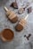Two chocolate-covered ice cream bars with wooden sticks lie on a textured gray surface. They are surrounded by chocolate drizzle and splatters, a spoon with melted chocolate, and several pieces of broken chocolate. A small glass cup filled with thick chocolate sauce is also present nearby.