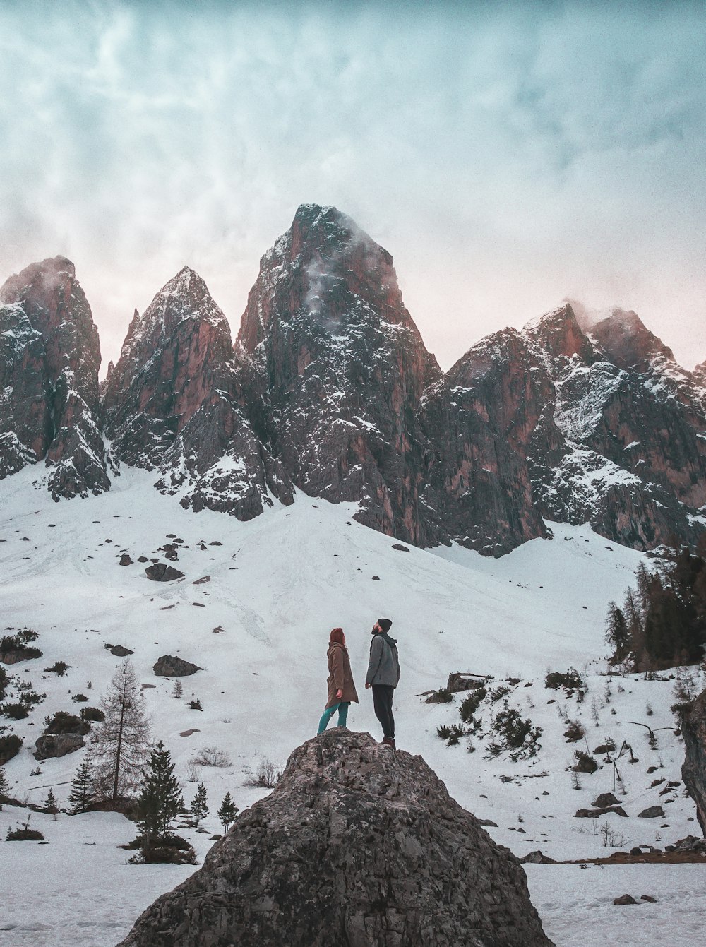 man and woman standing on the tip of a mountain with glacier mountains at the distance during day