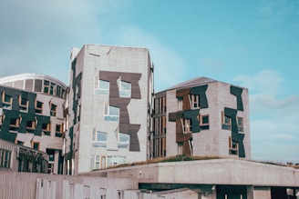 Modern architecture featuring several interconnected buildings with unique, angular window designs. The structures are composed primarily of stone with dark geometric accents and patches of vegetation on the roofs. The sky is clear with a few clouds visible.