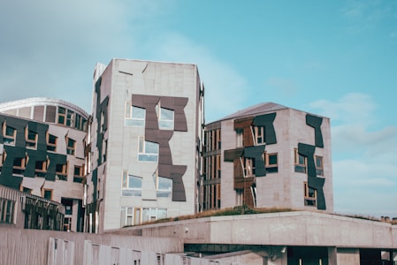 Modern architecture featuring several interconnected buildings with unique, angular window designs. The structures are composed primarily of stone with dark geometric accents and patches of vegetation on the roofs. The sky is clear with a few clouds visible.