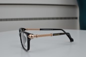 A pair of black eyeglasses with intricately designed gold-colored metal arms, placed on a smooth, white surface. The design features braided patterns, and the brand name is visible on the arm. The background consists of a minimalist, blurred setting with subtle horizontal lines.