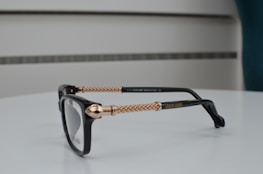 A pair of black eyeglasses with intricately designed gold-colored metal arms, placed on a smooth, white surface. The design features braided patterns, and the brand name is visible on the arm. The background consists of a minimalist, blurred setting with subtle horizontal lines.