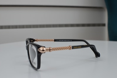 A pair of black eyeglasses with intricately designed gold-colored metal arms, placed on a smooth, white surface. The design features braided patterns, and the brand name is visible on the arm. The background consists of a minimalist, blurred setting with subtle horizontal lines.