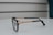 A pair of black eyeglasses with intricately designed gold-colored metal arms, placed on a smooth, white surface. The design features braided patterns, and the brand name is visible on the arm. The background consists of a minimalist, blurred setting with subtle horizontal lines.
