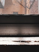 A close-up view of a metallic mesh screen with a background of orange brick walls. The image features strong contrasts between the structured lines of the mesh and the blurred, textured appearance of the bricks behind. The foreground includes a horizontal surface with a rough texture, partially illuminated by light.