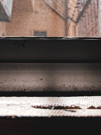 A close-up view of a metallic mesh screen with a background of orange brick walls. The image features strong contrasts between the structured lines of the mesh and the blurred, textured appearance of the bricks behind. The foreground includes a horizontal surface with a rough texture, partially illuminated by light.