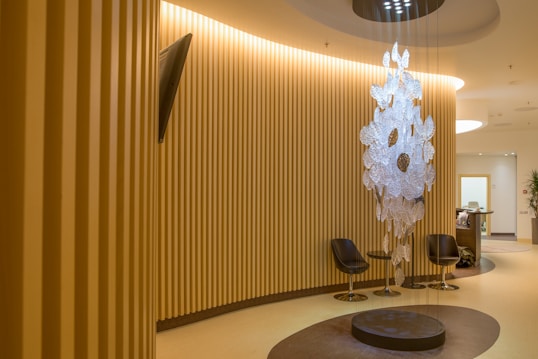 A modern, well-lit interior space with vertical wooden slats lining the walls, creating a warm and inviting atmosphere. A unique, artistic chandelier made of translucent materials hangs from the ceiling in the center, casting interesting shadows on the floor. Two sleek, black chairs are arranged around a circular, dark brown rug, while a small, contemporary reception desk can be seen in the background.