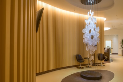 A modern, well-lit interior space with vertical wooden slats lining the walls, creating a warm and inviting atmosphere. A unique, artistic chandelier made of translucent materials hangs from the ceiling in the center, casting interesting shadows on the floor. Two sleek, black chairs are arranged around a circular, dark brown rug, while a small, contemporary reception desk can be seen in the background.