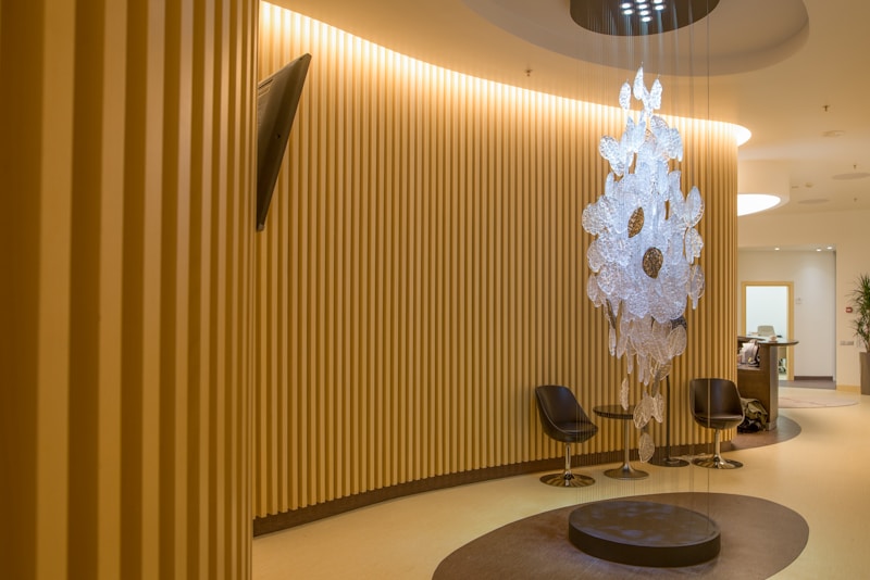 A modern, well-lit interior space with vertical wooden slats lining the walls, creating a warm and inviting atmosphere. A unique, artistic chandelier made of translucent materials hangs from the ceiling in the center, casting interesting shadows on the floor. Two sleek, black chairs are arranged around a circular, dark brown rug, while a small, contemporary reception desk can be seen in the background.