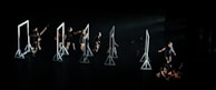 A group of dancers on a dark stage interacting with several geometric frames, each frame is lit emphasizing the performers' movements. The dancers are dressed in black, and their actions seem choreographed in a dynamic and expressive manner.