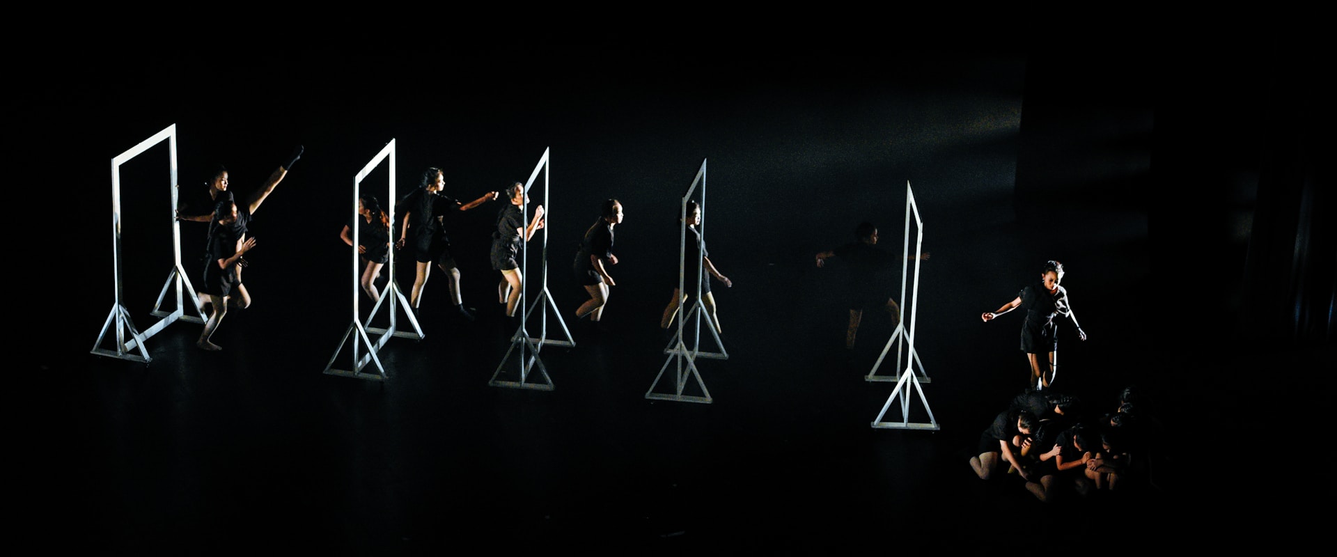 A group of dancers on a dark stage interacting with several geometric frames, each frame is lit emphasizing the performers' movements. The dancers are dressed in black, and their actions seem choreographed in a dynamic and expressive manner.