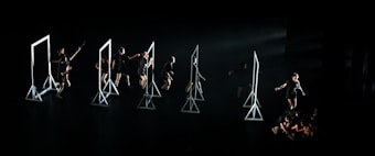 A group of dancers on a dark stage interacting with several geometric frames, each frame is lit emphasizing the performers' movements. The dancers are dressed in black, and their actions seem choreographed in a dynamic and expressive manner.
