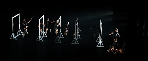 A group of dancers on a dark stage interacting with several geometric frames, each frame is lit emphasizing the performers' movements. The dancers are dressed in black, and their actions seem choreographed in a dynamic and expressive manner.