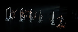 A group of dancers on a dark stage interacting with several geometric frames, each frame is lit emphasizing the performers' movements. The dancers are dressed in black, and their actions seem choreographed in a dynamic and expressive manner.