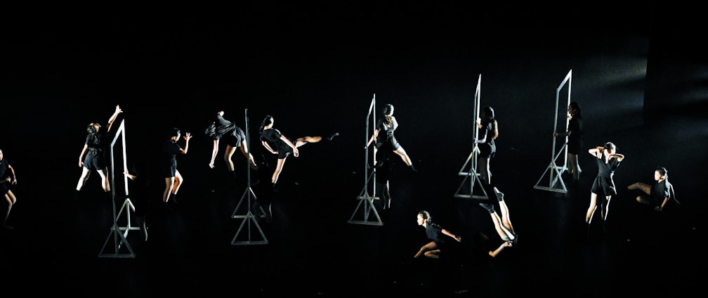 A group of individuals dressed in black performs a synchronized dance routine on a dark stage. Geometric frames are placed as props, with dancers interacting with them in various poses and movements.