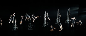 A group of individuals dressed in black performs a synchronized dance routine on a dark stage. Geometric frames are placed as props, with dancers interacting with them in various poses and movements.