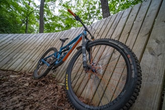 A mountain bike is propped against a wooden slatted wall in a forested area. The bike features a blue and orange frame with thick tires designed for off-road use. The ground is covered with dry leaves, and tall trees are visible in the background.