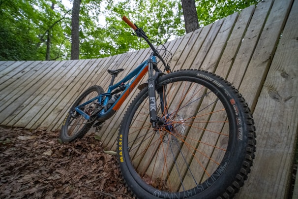 A mountain bike is propped against a wooden slatted wall in a forested area. The bike features a blue and orange frame with thick tires designed for off-road use. The ground is covered with dry leaves, and tall trees are visible in the background.