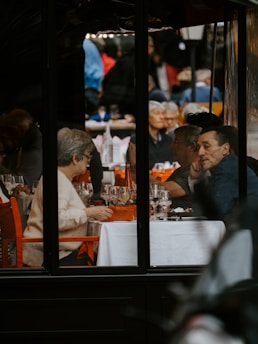 A group of elderly people sitting around a table in a restaurant, engaged in conversation and enjoying their meal. The atmosphere appears lively with many patrons visible in the background. The setting is warm with tables adorned with white tablecloths and orange accents.
