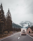 A camper van drives along a road flanked by tall evergreen trees, heading towards a fog-covered mountain range. Two bicycles are mounted on the back of the van. On the right side of the road, there is a small chapel with a green roof and a cross on top.