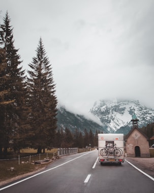 A camper van drives along a road flanked by tall evergreen trees, heading towards a fog-covered mountain range. Two bicycles are mounted on the back of the van. On the right side of the road, there is a small chapel with a green roof and a cross on top.