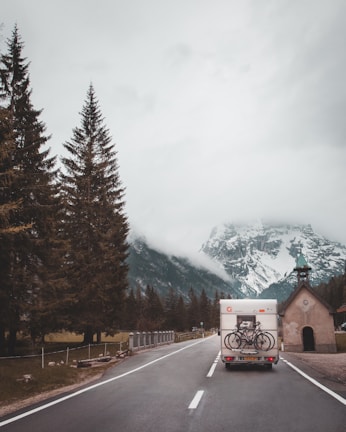 A camper van drives along a road flanked by tall evergreen trees, heading towards a fog-covered mountain range. Two bicycles are mounted on the back of the van. On the right side of the road, there is a small chapel with a green roof and a cross on top.