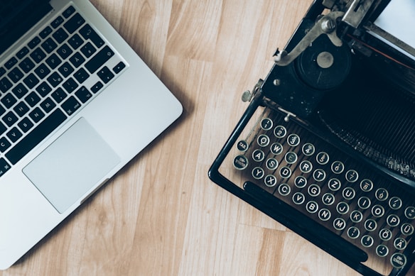 A modern laptop placed on a wooden surface beside a vintage typewriter, highlighting the contrast between old and new technology.