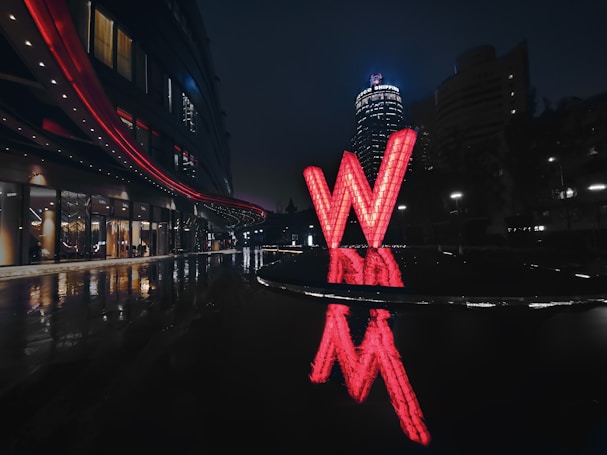 A large illuminated red 'W' structure stands prominently in an outdoor urban environment, reflecting on the wet ground. The surrounding area is dark, highlighting the bright and colorful installation. A modern building with long rows of windows and red accent lighting is visible on the left, together with a city skyline in the background.