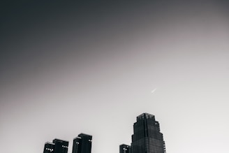 A skyline with a prominent modern skyscraper set against a gradient sky that transitions from dark grey to light grey. There is a linear object, possibly an airplane or shooting star, visible in the upper portion of the sky.