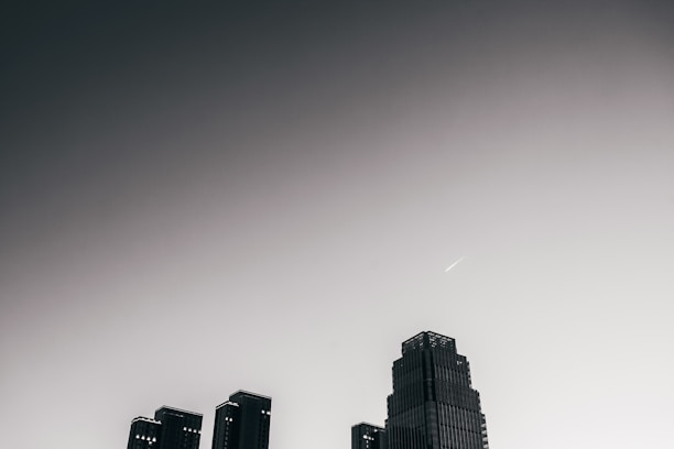 A skyline with a prominent modern skyscraper set against a gradient sky that transitions from dark grey to light grey. There is a linear object, possibly an airplane or shooting star, visible in the upper portion of the sky.