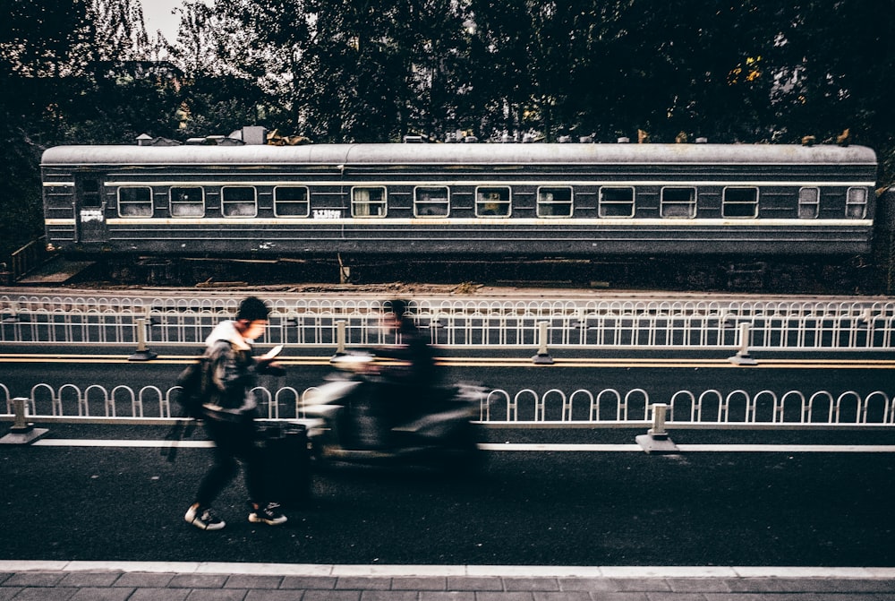 person walking on road near another person riding motorcycle beside train and trees