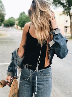 A person stands outdoors holding a skateboard. They are dressed in a black top and blue jeans with a denim jacket draped around their shoulders. Their blonde hair is loose, and they are listening to music through earbuds.