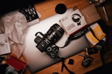 A camera with a lens is resting on a closed laptop amid a cluttered desk. There is a lens adapter box nearby, along with various camera accessories, papers, a lens cap, and scattered items like keys and cables.