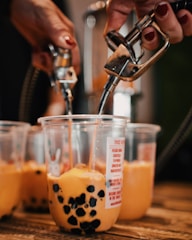 Hands are filling clear plastic cups with a brownish-orange liquid containing black tapioca pearls using metal dispensers. The cups are placed on a wooden surface and include various labels and graphics.