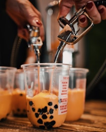 Hands are filling clear plastic cups with a brownish-orange liquid containing black tapioca pearls using metal dispensers. The cups are placed on a wooden surface and include various labels and graphics.