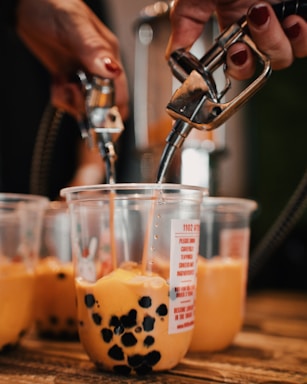 Hands are filling clear plastic cups with a brownish-orange liquid containing black tapioca pearls using metal dispensers. The cups are placed on a wooden surface and include various labels and graphics.