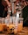 Hands are filling clear plastic cups with a brownish-orange liquid containing black tapioca pearls using metal dispensers. The cups are placed on a wooden surface and include various labels and graphics.