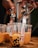 Hands are filling clear plastic cups with a brownish-orange liquid containing black tapioca pearls using metal dispensers. The cups are placed on a wooden surface and include various labels and graphics.