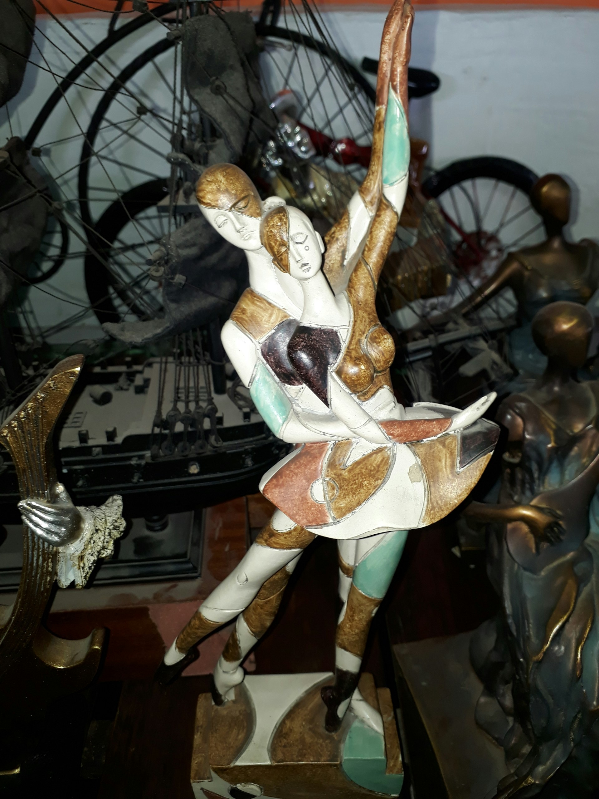 A stylized sculpture of a dancing couple depicted in abstract form, with exaggerated limbs and artistic detailing. The figures display grace and movement, captured mid-dance with one arm elegantly raised. The background includes various mechanical parts, possibly of bicycles, adding an industrial ambiance.