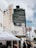 A street scene featuring a vintage theater marquee at the El Rey, displaying information for Zoe Church LA. People mill about under white tents that suggest an event or market. The background shows a tall black office building with the number 5455 and palm trees lining the street.