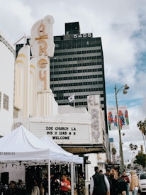 A street scene featuring a vintage theater marquee at the El Rey, displaying information for Zoe Church LA. People mill about under white tents that suggest an event or market. The background shows a tall black office building with the number 5455 and palm trees lining the street.