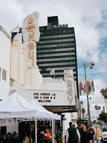 A street scene featuring a vintage theater marquee at the El Rey, displaying information for Zoe Church LA. People mill about under white tents that suggest an event or market. The background shows a tall black office building with the number 5455 and palm trees lining the street.